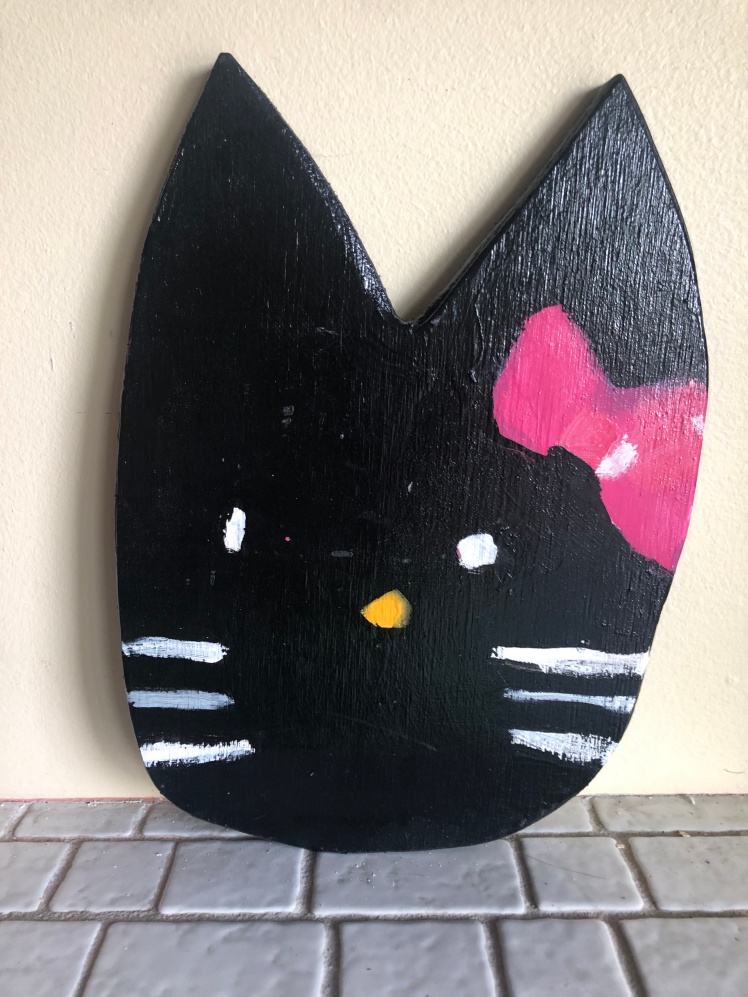 This Anthony Lister artwork was painted on plywood in 2010 and is a black kitty with white eyes and whiskers as well as a pink bow.
